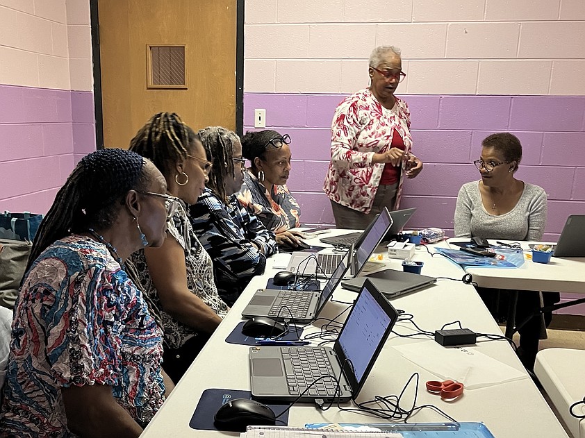 Five women sit around a table with laptops in front of them at a Tech Goes Home class while one trainer stands among them talking.